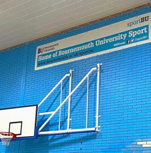 Bournemouth University's sports hall used by Sport BU - picture shows a basketball hoop on a wall mount, on a blue brick wall with 'Home of Bournemouth University Sport' above it