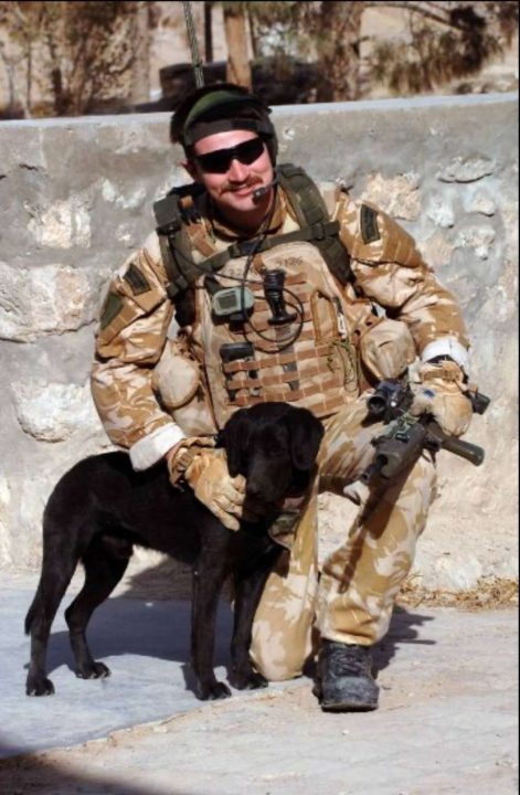 John Thornton in army gear, petting a black dog.