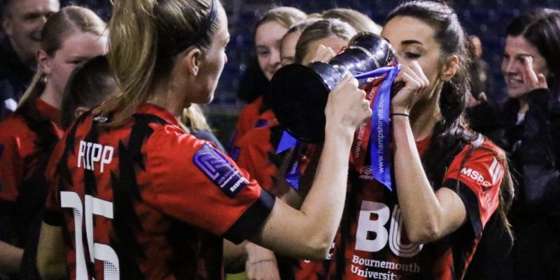 Kelly Fripp helping Maisy Smith drink out of the Hampshire FA Cup. They are both wearing the Bournemouth home shirt.