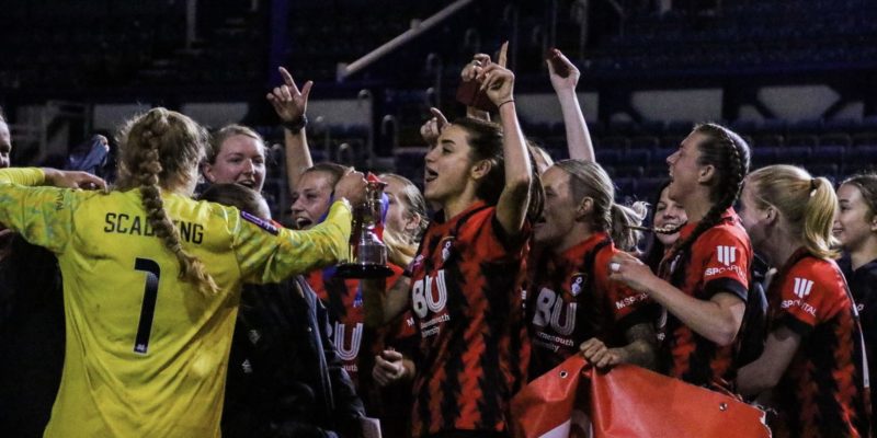 The Bournemouth team celebrate winning the Hampshire FA Cup. Maisy Smith is front and centre holding the trophy. The team is jumping up and down.