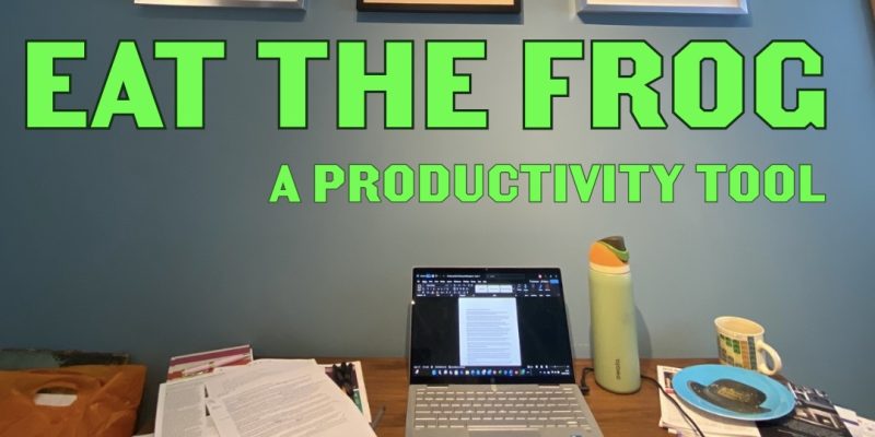 a cluttered desk with picture frames hanging on a blue wall, words 'EAT THE FROG: A productivity tool' written over the image in green.